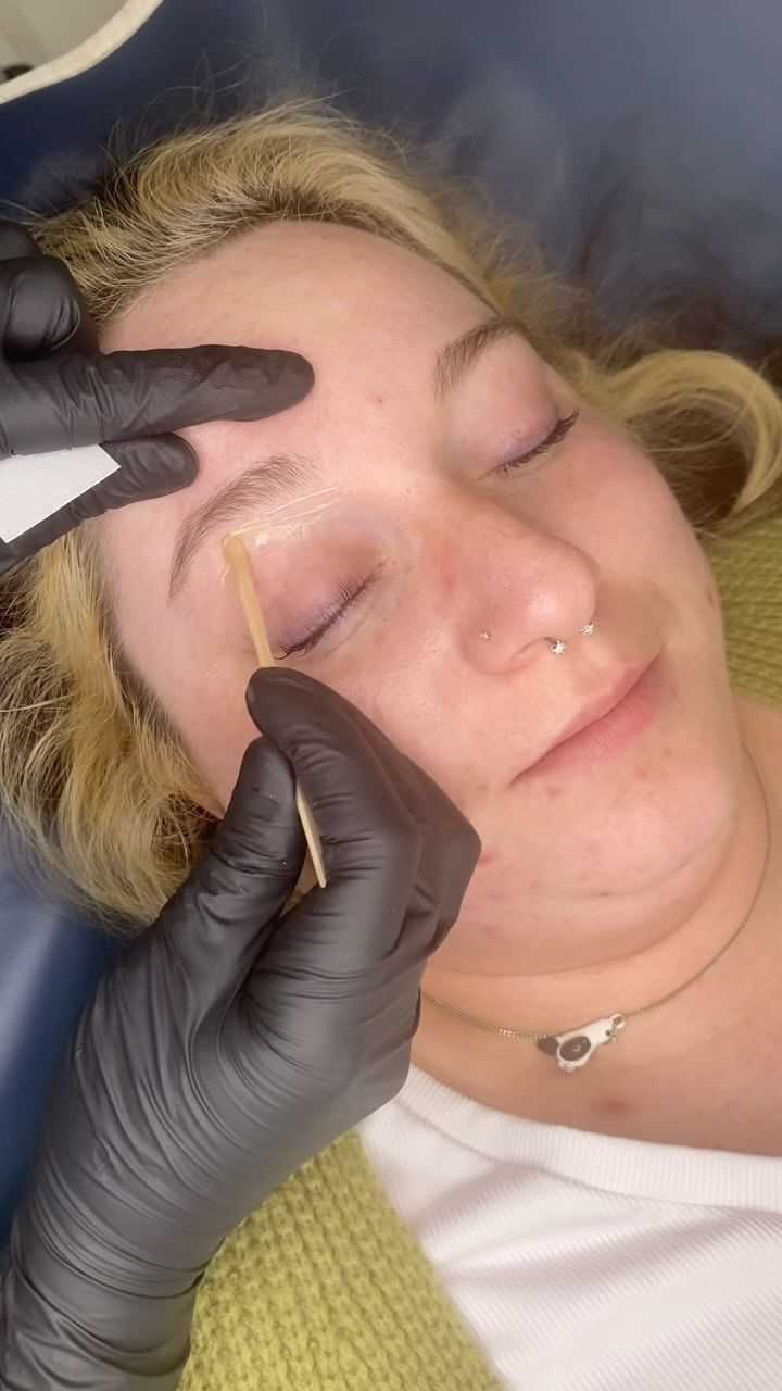 Person receiving eyebrow grooming with wooden applicator, eyes closed, relaxed expression.