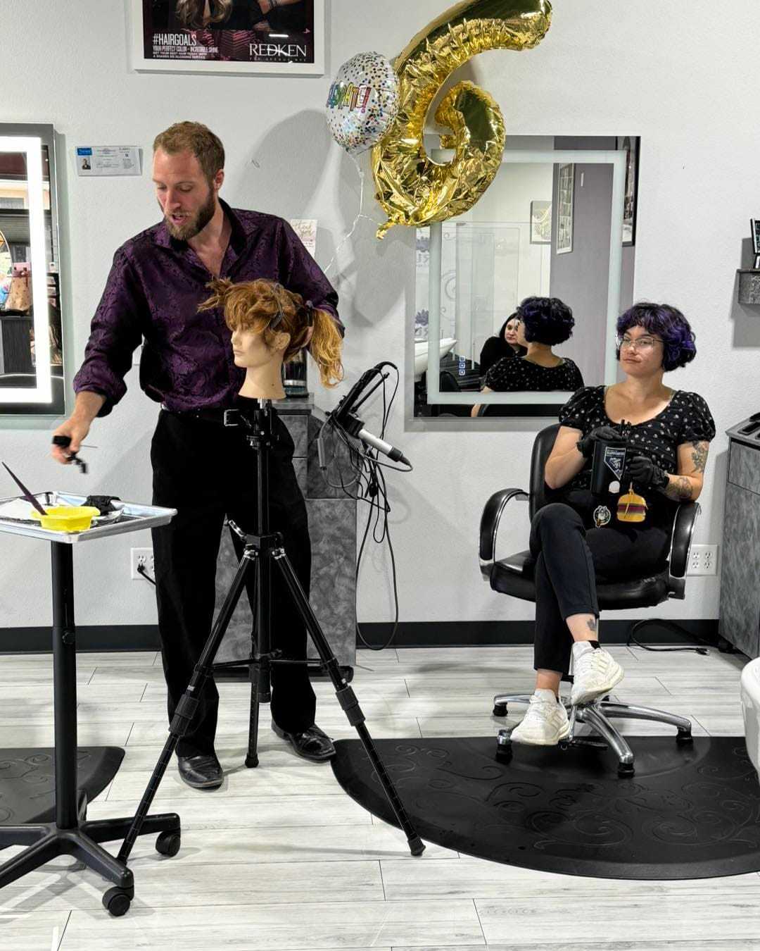 Hair stylist working with mannequin, woman seated with mirror reflection, balloons in background.