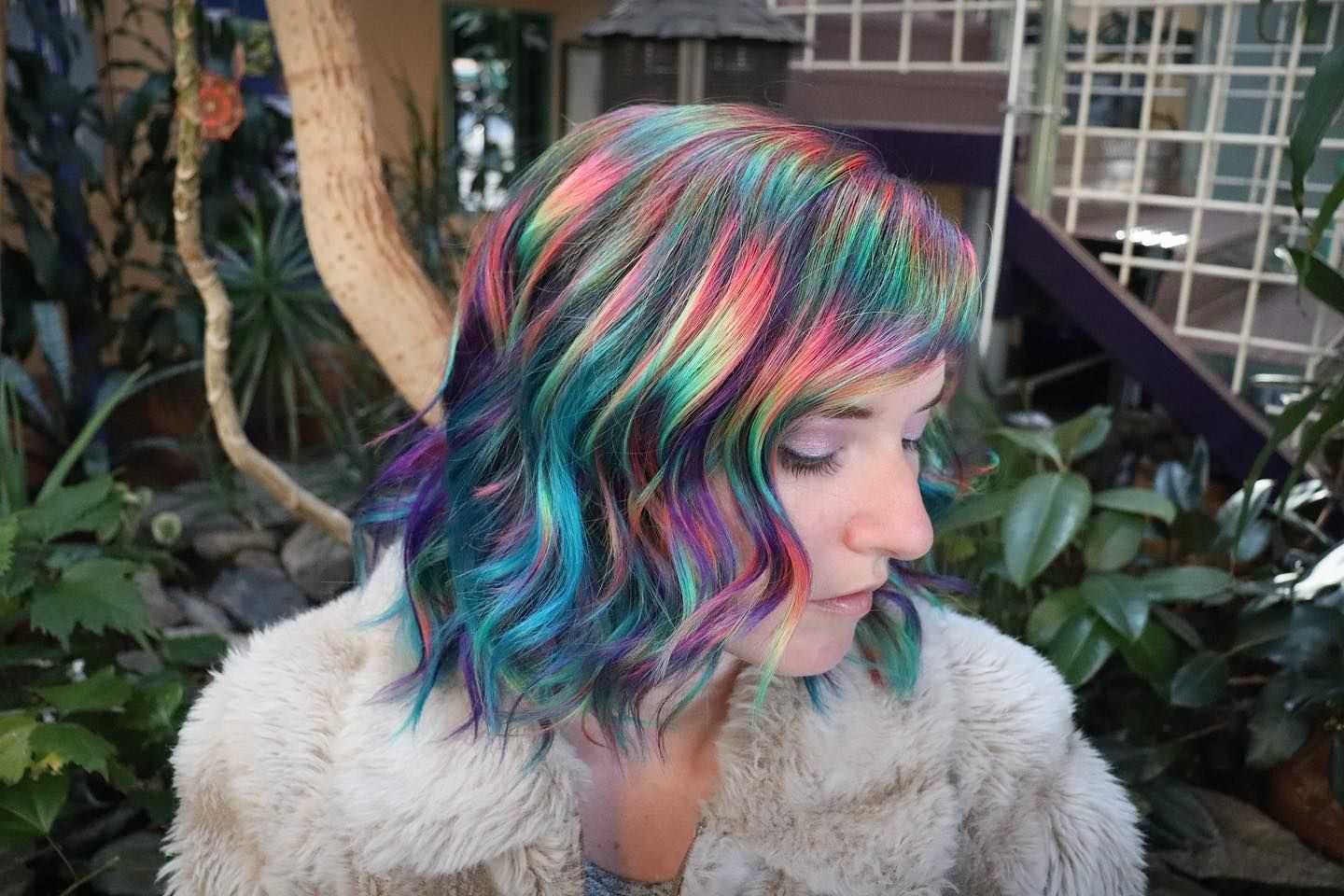 Woman with vibrant, multicolored hair in a lush, plant-filled setting.